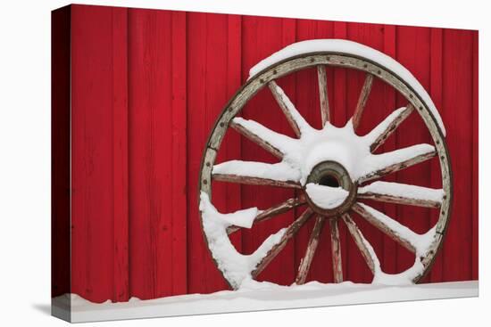 Snow-covered wagon wheels against red barn near town of Banff, Canadian Rockies, Alberta, Canada-Stuart Westmorland-Premier Image Canvas