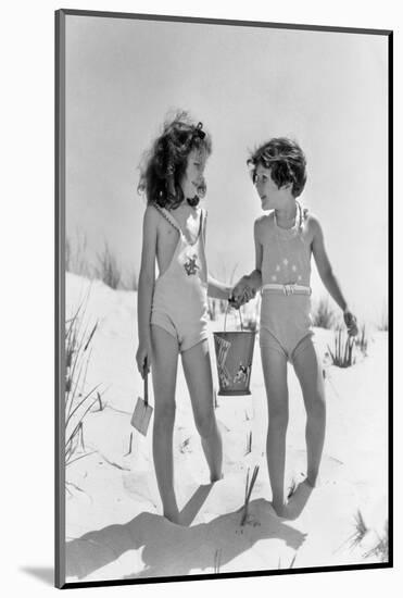 1930s TWO SMILING GIRLS SISTERS FRIENDS HOLDING HANDS AND SAND PAIL AND SHOVEL WALKING ON SAND DUNE-H. Armstrong Roberts-Mounted Photographic Print