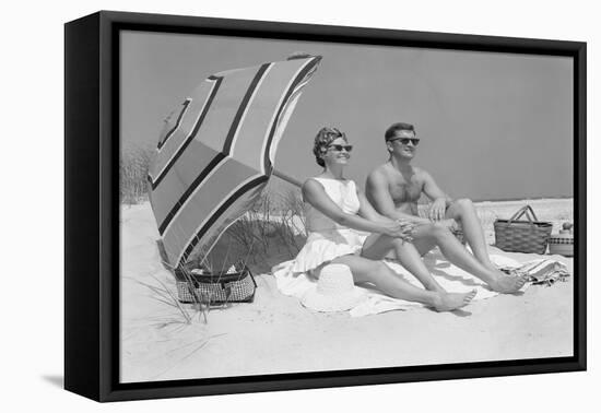 1960s COUPLE IN SUNGLASSES SITTING ON BEACH BLANKET WITH LEGS EXTENDED WITH UMBRELLA-H. Armstrong Roberts-Framed Premier Image Canvas