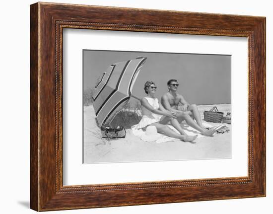 1960s COUPLE IN SUNGLASSES SITTING ON BEACH BLANKET WITH LEGS EXTENDED WITH UMBRELLA-H. Armstrong Roberts-Framed Photographic Print