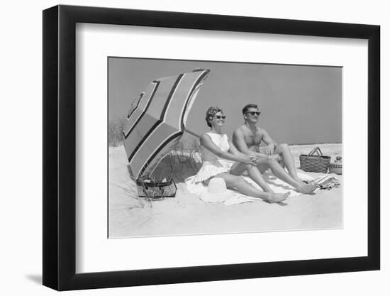 1960s COUPLE IN SUNGLASSES SITTING ON BEACH BLANKET WITH LEGS EXTENDED WITH UMBRELLA-H. Armstrong Roberts-Framed Photographic Print