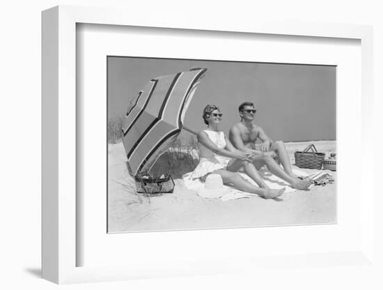 1960s COUPLE IN SUNGLASSES SITTING ON BEACH BLANKET WITH LEGS EXTENDED WITH UMBRELLA-H. Armstrong Roberts-Framed Photographic Print