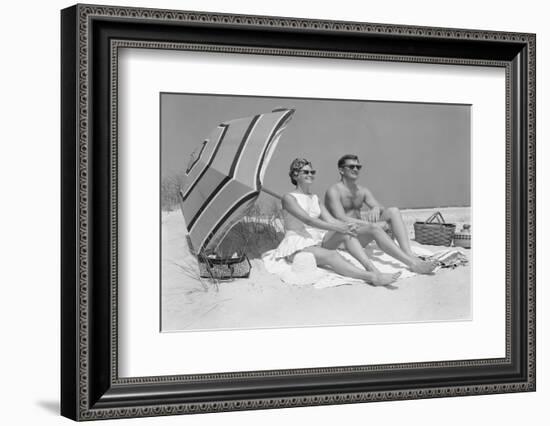 1960s COUPLE IN SUNGLASSES SITTING ON BEACH BLANKET WITH LEGS EXTENDED WITH UMBRELLA-H. Armstrong Roberts-Framed Photographic Print