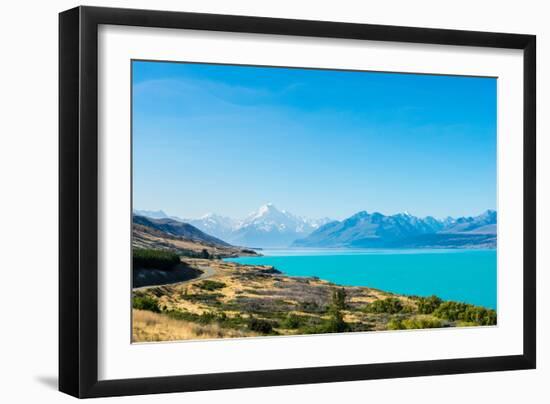 A road winds along the edge of a turquoise blue lake with mountains in the distance, New Zealand-Logan Brown-Framed Photographic Print