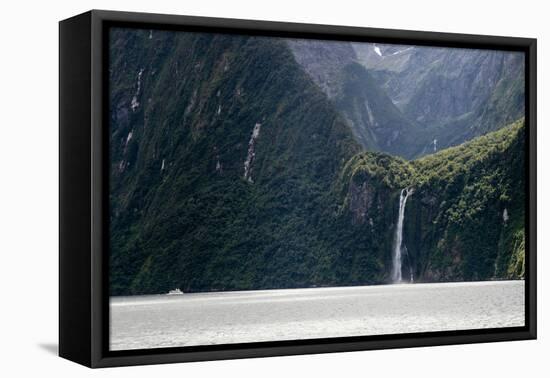 A sightseeing ship dwarfed by a tall waterfall in a fjord, South Island, New Zealand, Pacific-Logan Brown-Framed Premier Image Canvas