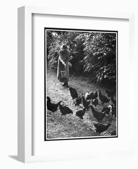 An Older Woman in a Long Dress and Wide-Brimmed Hat Throws Handfuls of Chicken Feed-null-Framed Art Print