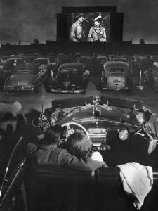 Young Couple Snuggling in Convertible as They Watch Large Screen Action at a Drive-In Movie Theater