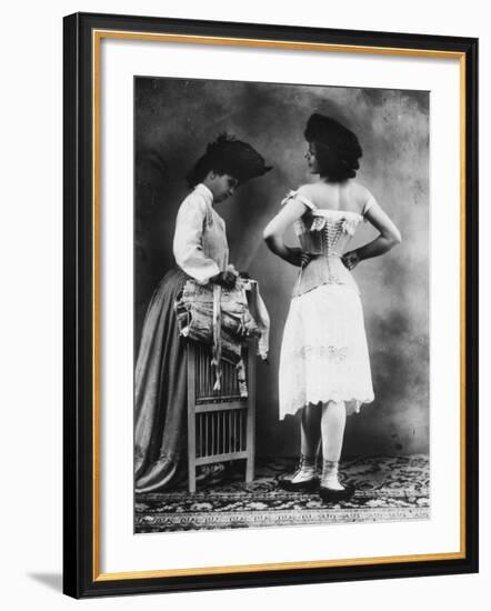 Lady Tries on a Corset While Another Woman Waits with Another One--Framed Photographic Print