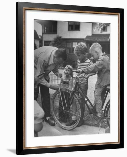 Man Fixing Basket on Bicycle as Children Watch Attentively-Nina Leen-Framed Photographic Print