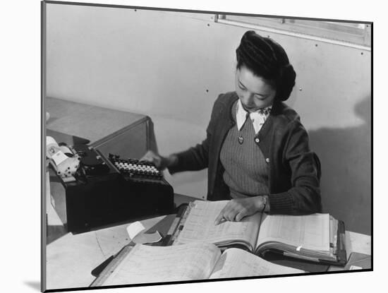 Mrs Teruko Kiyomura, bookkeeper, seated at desk, operating an adding machine while reading a ledger-Ansel Adams-Mounted Photographic Print