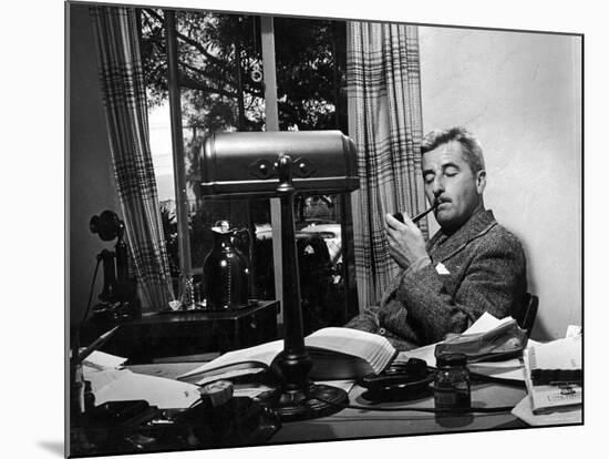 Novelist and Script Writer William Faulkner Smoking a Pipe at His Desk at Warner Brothers Studios-null-Mounted Premium Photographic Print