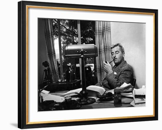 Novelist and Script Writer William Faulkner Smoking a Pipe at His Desk at Warner Brothers Studios-null-Framed Premium Photographic Print