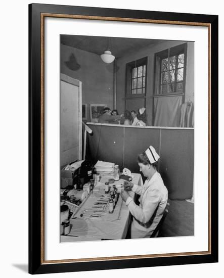 Nurse Sitting at Table with Medical Supplies While Doctors Examine Patient in Background-Wallace Kirkland-Framed Premium Photographic Print
