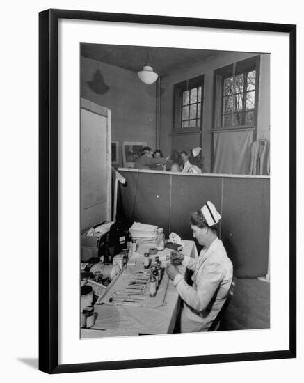 Nurse Sitting at Table with Medical Supplies While Doctors Examine Patient in Background-Wallace Kirkland-Framed Premium Photographic Print