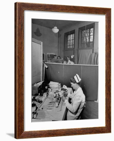 Nurse Sitting at Table with Medical Supplies While Doctors Examine Patient in Background-Wallace Kirkland-Framed Premium Photographic Print