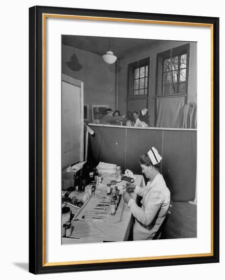 Nurse Sitting at Table with Medical Supplies While Doctors Examine Patient in Background-Wallace Kirkland-Framed Premium Photographic Print