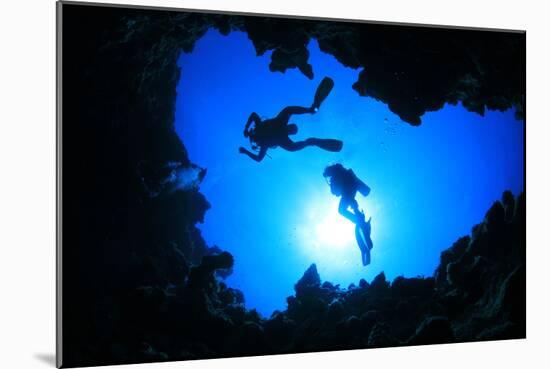 Scuba Divers Descend into an Underwater Cavern. Silhouettes against Sunburst-Rich Carey-Mounted Photographic Print